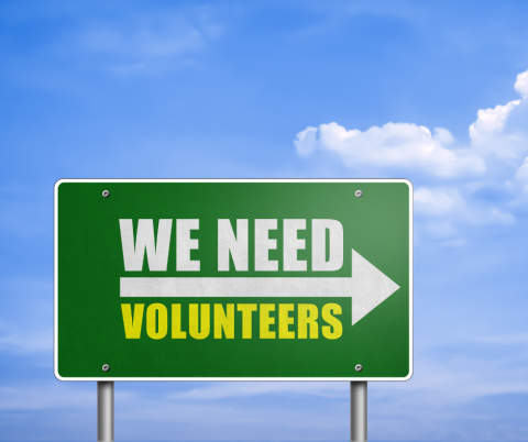 A green road sign that says, "We Need Volunteers", with an arrow pointing towards the right.  Set against a blue sky with a couple of clouds.