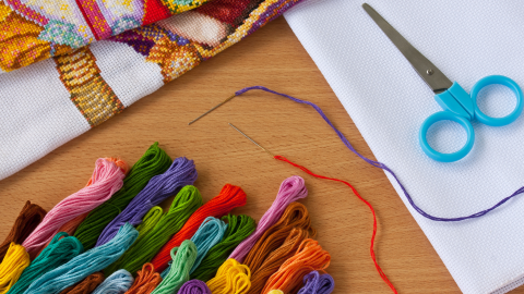 Supplies for cross stitch, spread out on a table.  Skeins of floss, needle and thread, scissors, and fabric.