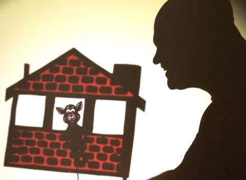 Shadow of a man on right side of picture. Features a brick house with three windows. A pig is seen looking through the middle window.