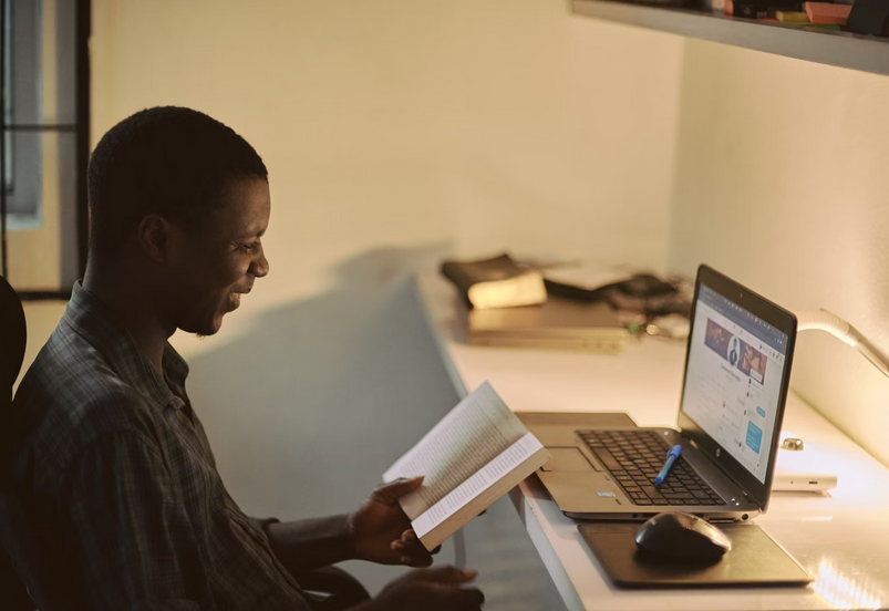 photo of someone smiling while reading a book at a desk in front a laptop 